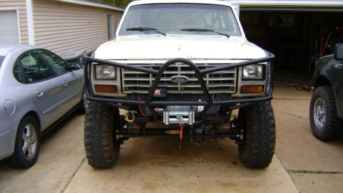 1986 Ford Bronco - Front view of a white 1980s Ford Bronco SUV lifted on large mud-terrain tires with a heavy tubular front bumper/brush guard and a mounted w…