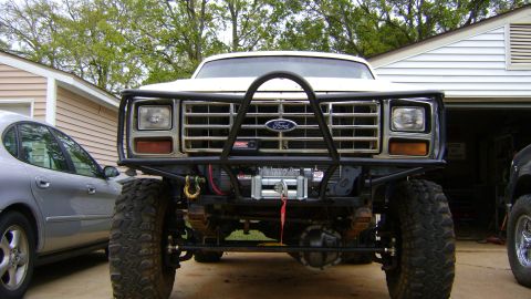 1986 Ford Bronco - Head-on view of a lifted white Ford Bronco in a driveway with a black tubular front bumper and full grille guard hoop, with a Warn winch mo…