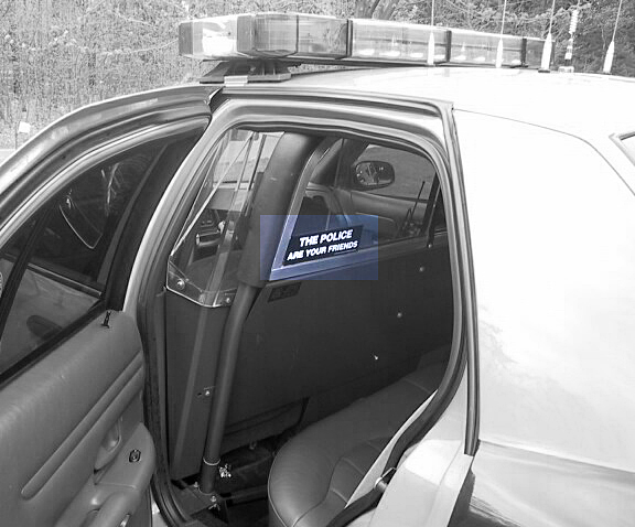 Interior of a police car, showing the back seat area with a partition and a sticker that says 'THE POLICE ARE YOUR FRIENDS'