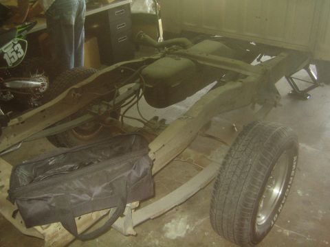 1990 Ford Bronco - bare truck frame rests on a garage floor showing a fuel tank and crossmembers, with a spare wheel, tool bag and a dirt bike alongside.