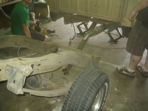 1990 Ford Bronco - truck frame and driveshaft are pulled out from under a raised tan cab as a man kneels working nearby on the garage floor.