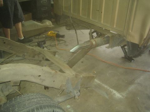 1990 Ford Bronco - Bare truck frame rails and a driveshaft lie on a garage floor beneath a raised tan cab supported on scissor jacks.
