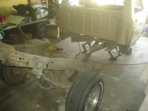 1990 Ford Bronco - tan pickup cab is raised on scissor jacks while its bare frame and rear wheel are pulled forward across the garage floor.