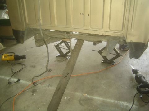 1990 Ford Bronco - tan vehicle cab sits raised on scissor jacks over a wooden beam, with reciprocating saws and an orange extension cord on the garage floor.