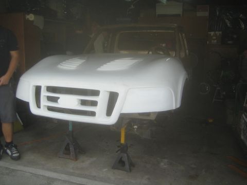 1990 Ford Bronco - white fiberglass one-piece front clip with hood, fenders and twin grille openings rests on jack stands in front of a tan project truck.