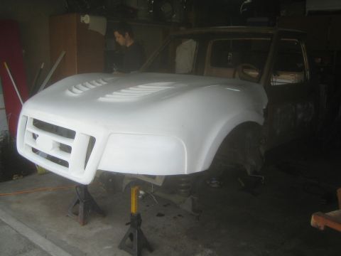 1990 Ford Bronco - white fiberglass front clip is test-fitted to a tan project truck on jack stands as a man works in the background.