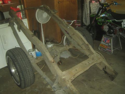 1990 Ford Bronco - bare ladder-type truck frame sits on its wheels in a garage next to a green dirt bike and a fan.