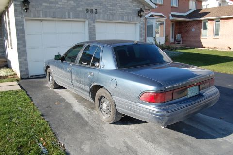 1985 Mercury Grand Marquis - Blue sedan parked in front of a residential garage. The car appears to be a 1990s model.