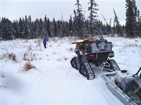 2000 Ford E350 - An ATV equipped with tracks tows a sled through a snow-covered field, with a person walking in the distance near a treeline.