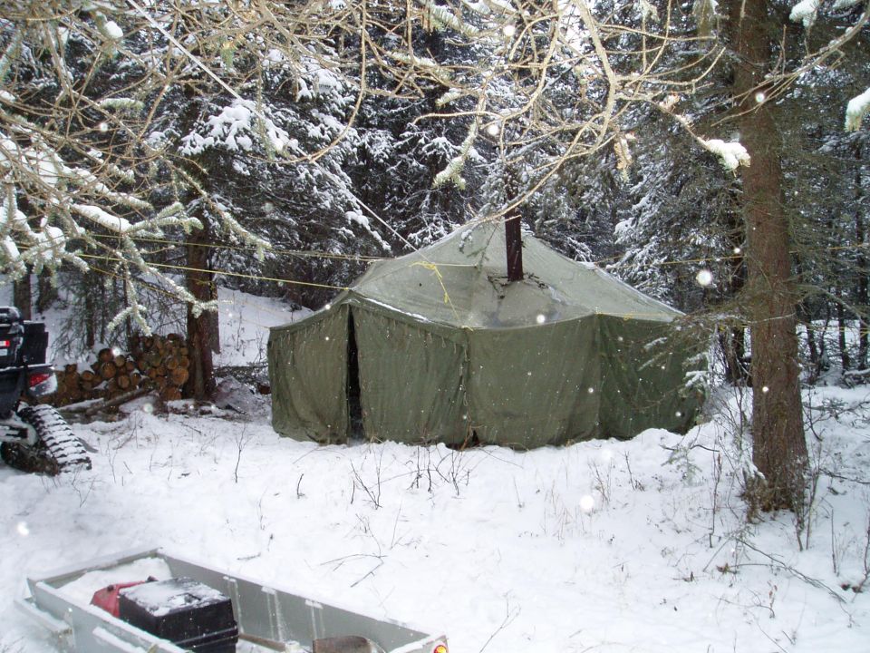 Tent set up in snowy woods with a trailer and tracked vehicle nearby