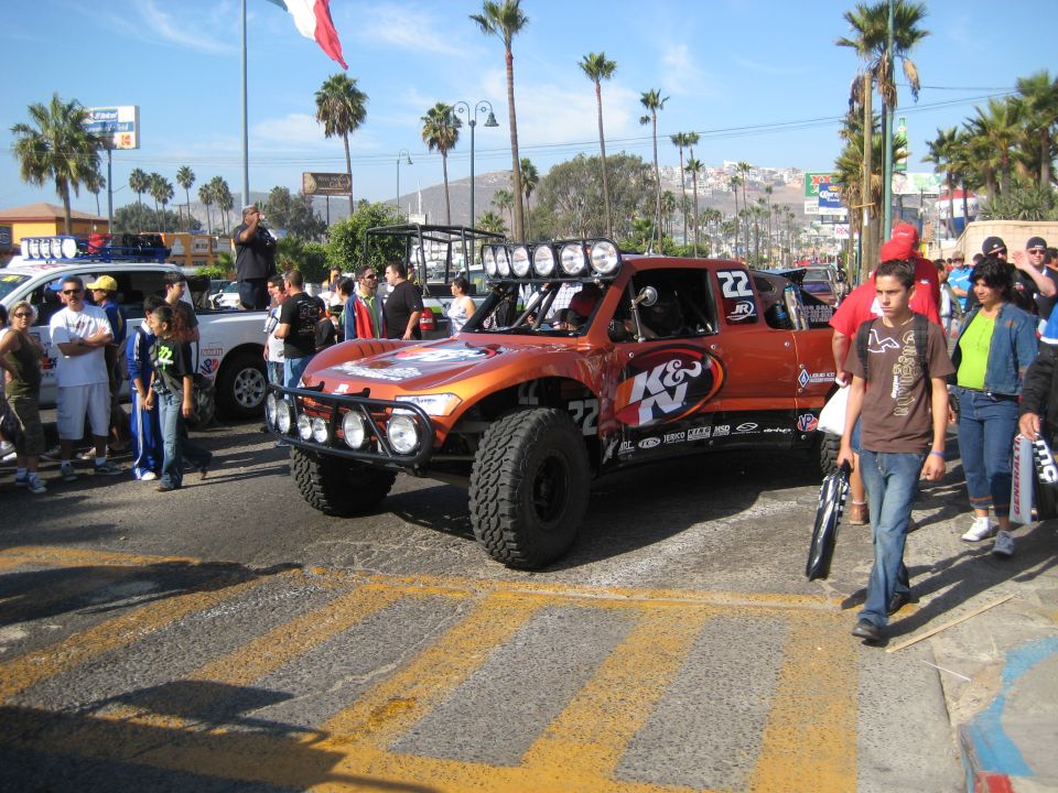 Orange off-road racing truck with K&N logos and number 22, parked on a street with people around. Another white truck is visible behind it