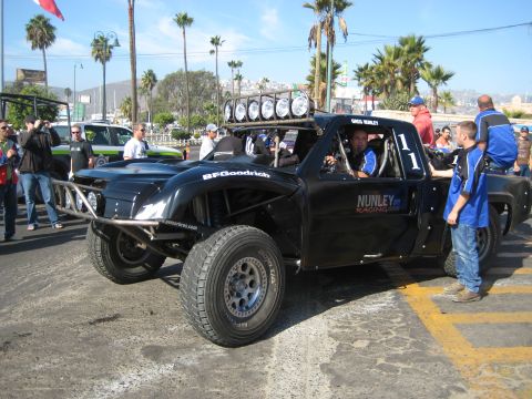 1996 Ford Bronco - A modified black truck with racing decals and multiple lights on the roof is parked on a street with people around.