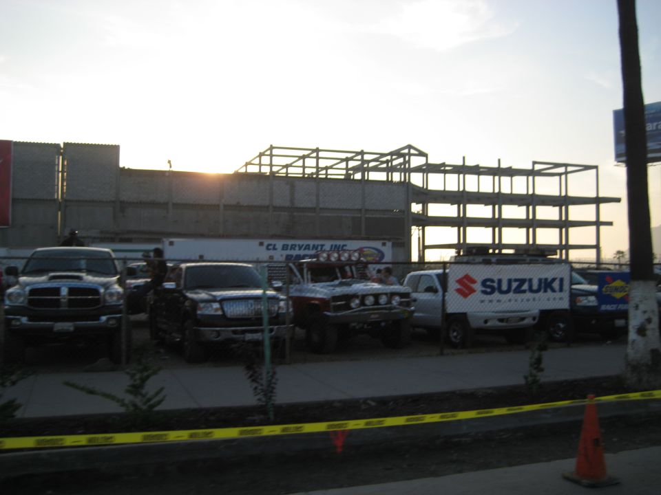 Several trucks and a Suzuki van are parked in front of a building under construction. A banner on the van reads SUZUKI