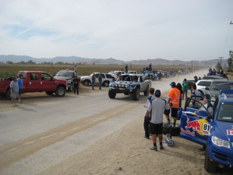 1996 Ford Bronco - A line of off-road race trucks and support vehicles are gathered on a dirt road, with spectators observing the scene.