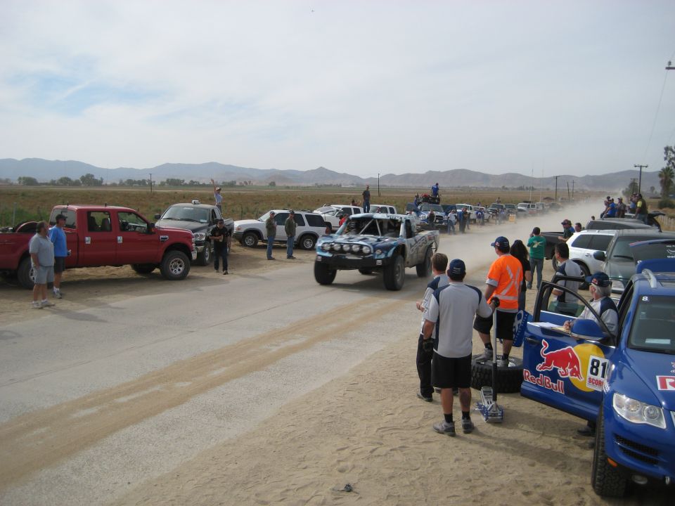 Off-road race trucks and support vehicles line a dirt road, with spectators watching