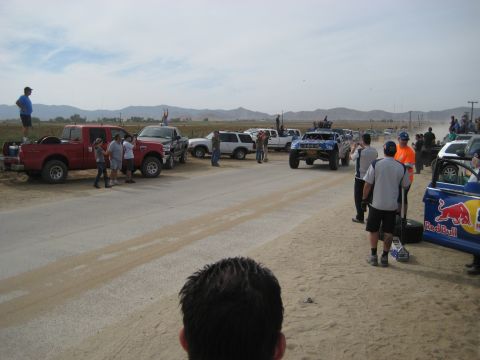 1996 Ford Bronco - A collection of trucks and off-road vehicles are staged on a dusty road, with people observing the scene.