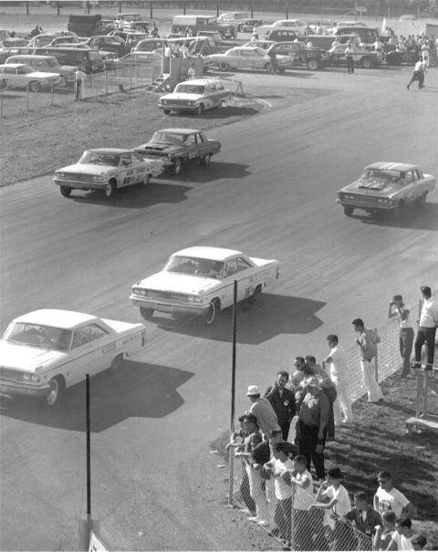 1963 Ford Galaxie - A field of stock cars, including a white 1963 Ford Galaxie, compete on a race track with spectators watching from the sidelines.