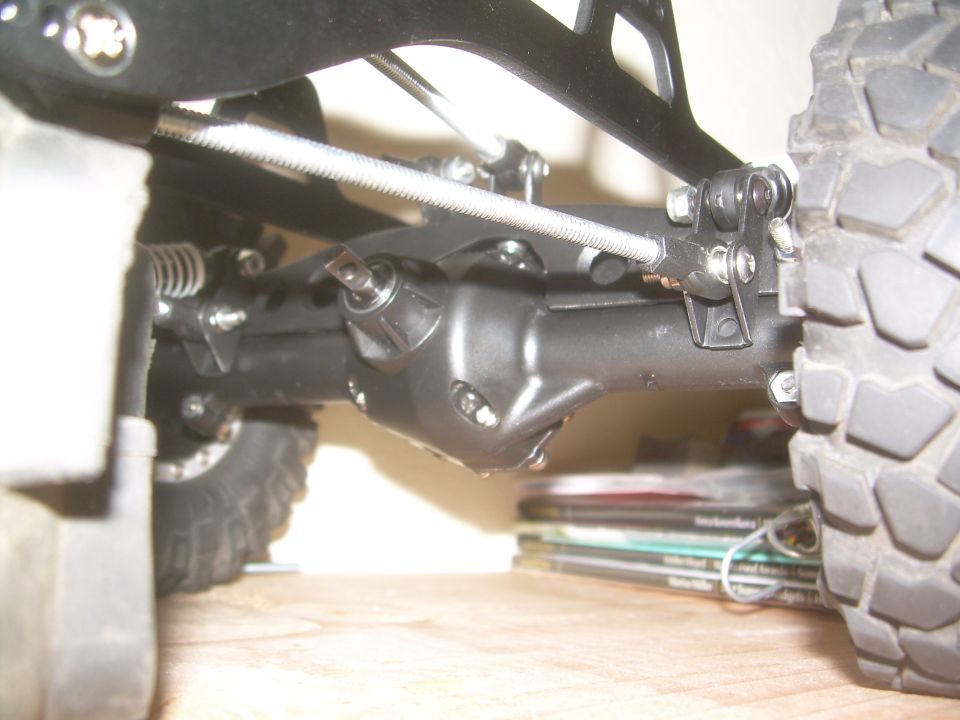 Close-up of a remote control car's rear axle, suspension components, and tire tread. Stack of magazines visible below