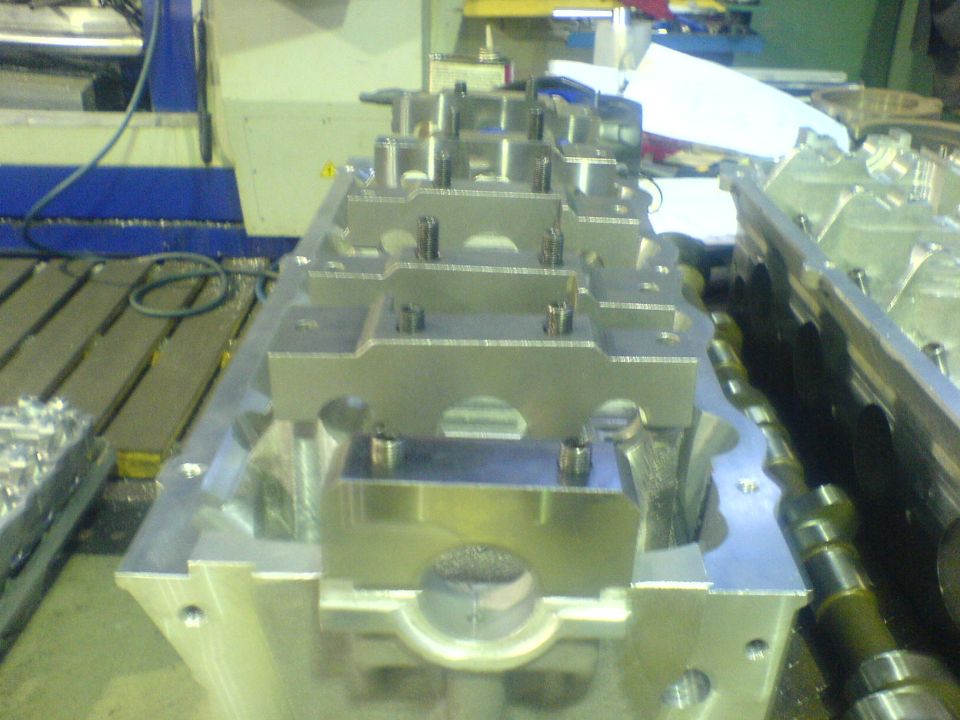 Machined aluminum cylinder head with cam tower bosses on a mill table, camshaft alongside