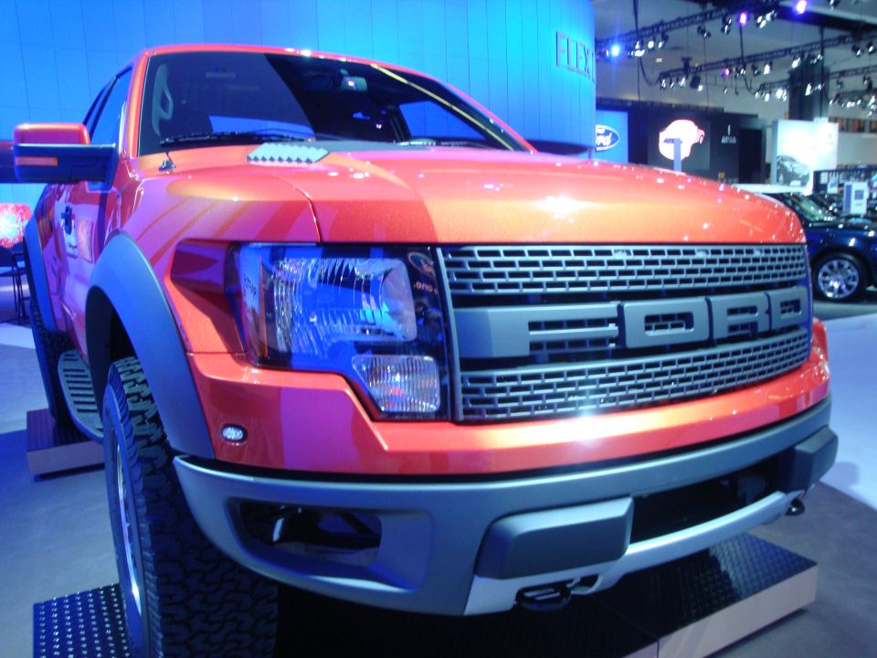 Orange Ford F-150 Raptor front end with large 'FORD' grille. Another car is visible in the background