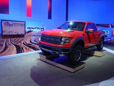 1993 Ford Taurus - A bright orange Ford SVT Raptor pickup truck is showcased at an auto show with a promotional video playing on a screen behind it.