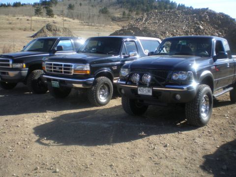 1996 Ford Bronco - Three black trucks and SUVs, including a 1996 Ford Bronco, are parked on a dirt lot with hills in the background.