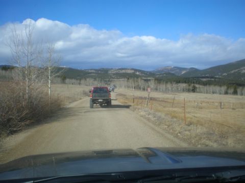1996 Ford Bronco - A red pickup truck drives down a dusty road with other vehicles ahead, surrounded by dry fields and distant mountains.