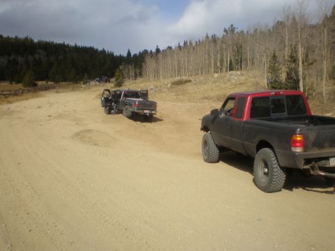 1996 Ford Bronco - Two pickup trucks are parked on a dirt road in a wooded area, with one truck in the foreground and another further back.