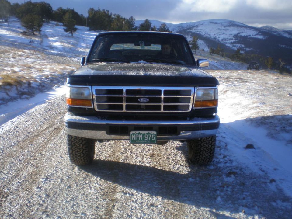 Black Ford Bronco on a snowy dirt road, front view. Chrome grille, aftermarket tires