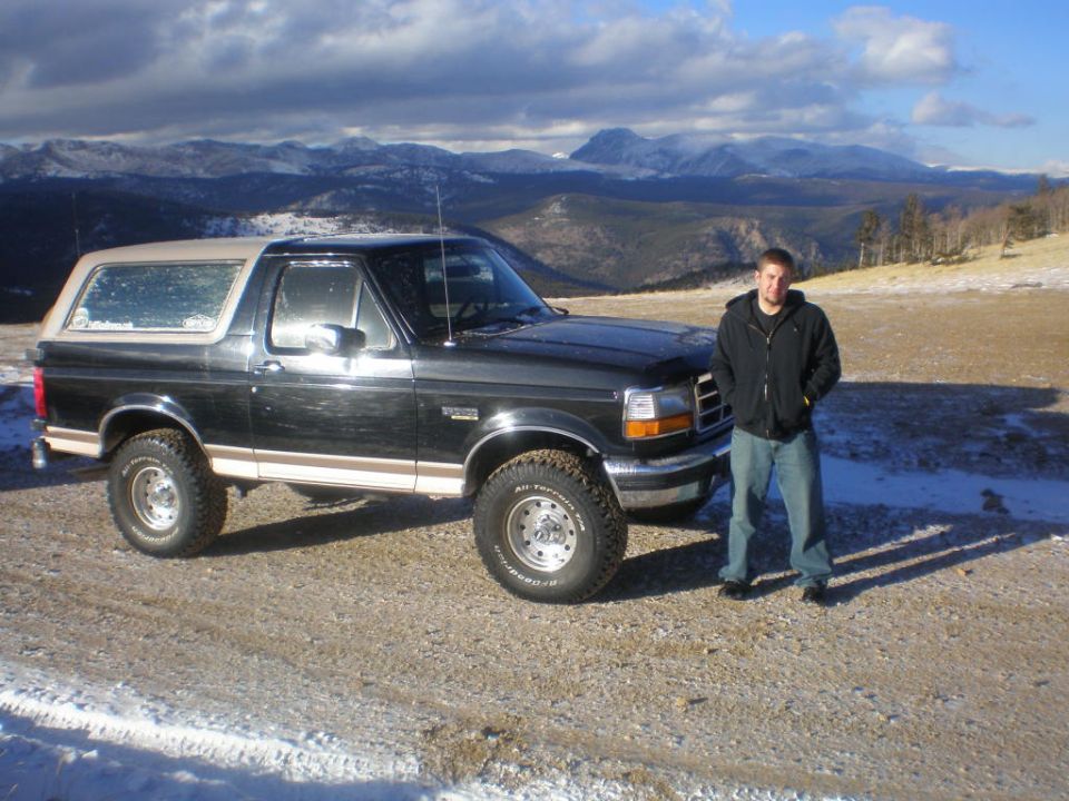 Black 1996 Ford Bronco with tan top parked on a dirt road with snow, mountains in background