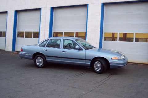 2001 Ford Crown Victoria - A light blue sedan is parked in front of a building with garage doors.