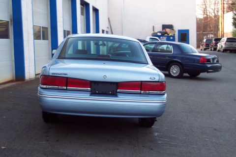 2001 Ford Crown Victoria - A light blue 2001 Ford Crown Victoria is parked in front of a building with a blue and white facade, with another Crown Victoria behind it.
