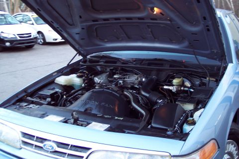 2001 Ford Crown Victoria - The engine bay of a 2001 Ford Crown Victoria is shown with the hood open. Two other vehicles are parked behind it.