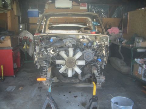 1990 Ford Bronco - head-on view shows a V-engine with a large white fan and coil-spring suspension mounted in a tan project truck on jack stands.
