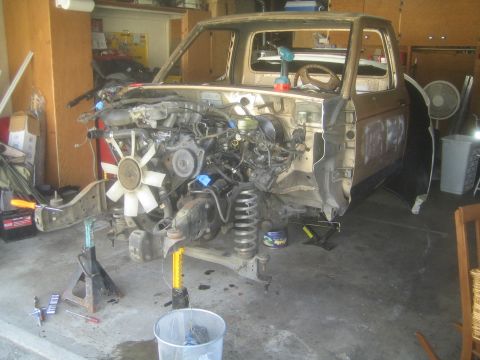 1990 Ford Bronco - side view shows a tan project truck on jack stands with its V-engine, white fan and front suspension installed but body front clip off.
