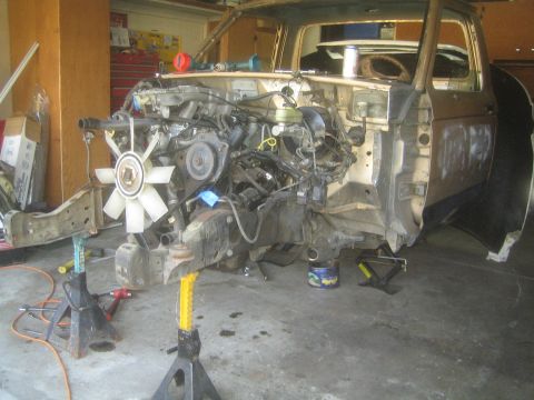 1990 Ford Bronco - partially disassembled pickup truck rests on jack stands in a cluttered garage, engine and frame rail exposed.