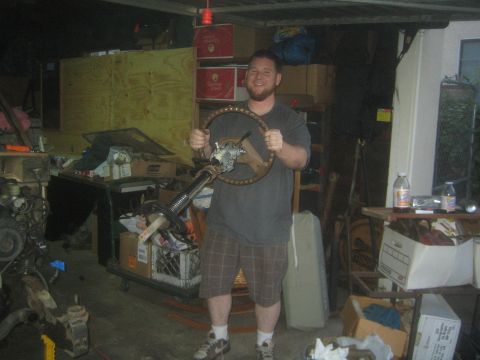 1990 Ford Bronco - bearded man in a gray shirt grins while holding a removed steering column and wheel in a cluttered garage.