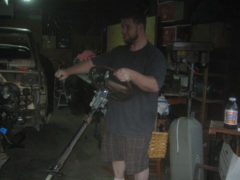 1990 Ford Bronco - man holds a long steering column and wheel assembly in a dimly lit, cluttered garage workshop.
