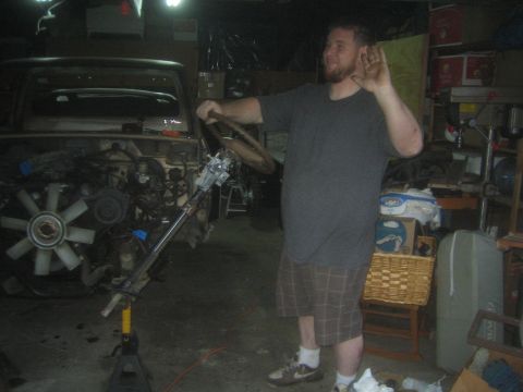 1990 Ford Bronco - man gestures with one hand while holding a steering column, standing beside a stripped truck in a dim garage.