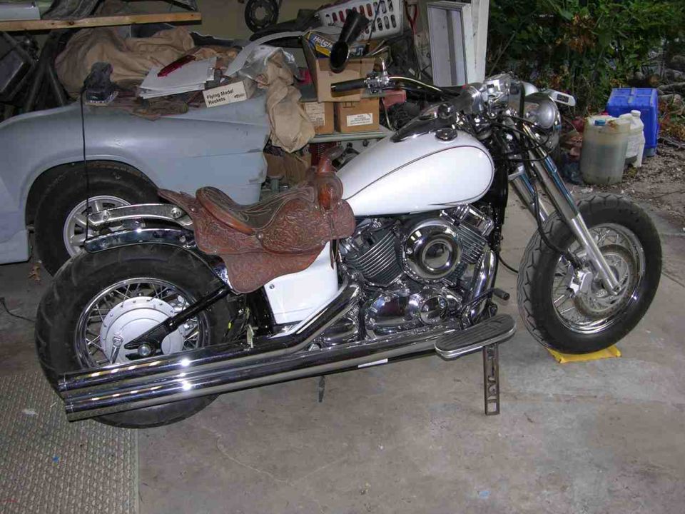 White Yamaha V Star cruiser with a leather horse saddle mounted as its seat, parked in a cluttered garage