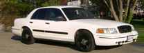 2003 Ford Crown Victoria - A white 2003 Ford Crown Victoria is parked in front of some trees.