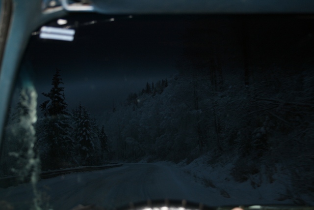 View from inside a vehicle looking out at a dark, snowy road winding through a forest at dusk