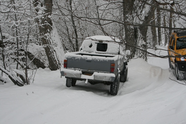 Gray pickup truck from rear, covered in snow, with a yellow SUV to the right