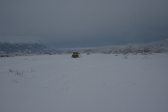 Yellow vehicle driving through a vast, snow-covered field under a cloudy sky with distant mountains