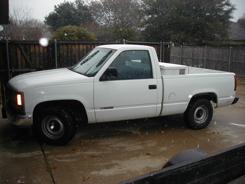 White regular-cab pickup with bed-mounted toolbox parked on wet driveway by a wooden fence