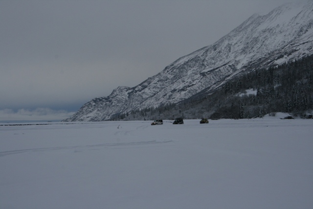Three vehicles parked on a vast, snow-covered plain with a large, snow-capped mountain in the background