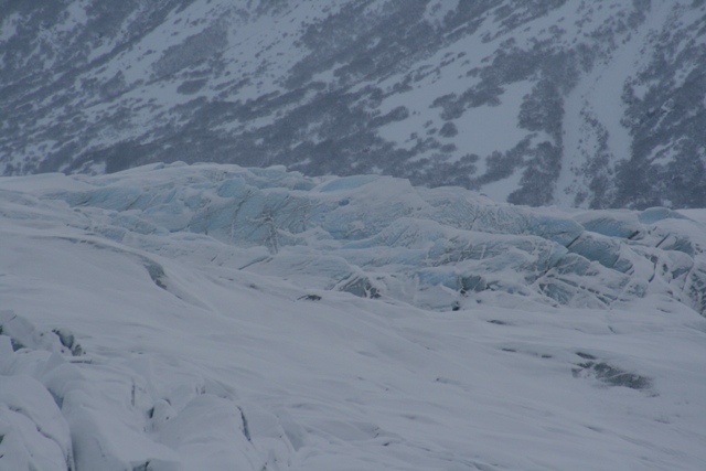 Snowy mountain slopes above a textured, blue-tinged glacier