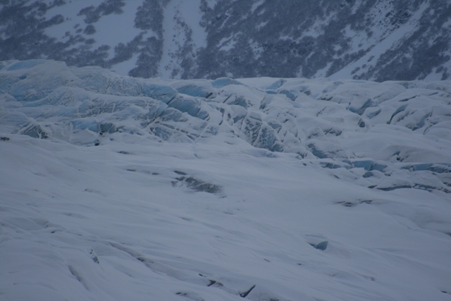 Snow-covered glacier with blue ice formations and a distant, snow-dusted mountain