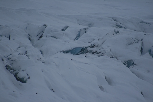 Snow-covered glacier surface with deep crevasses and blue ice visible in some cracks
