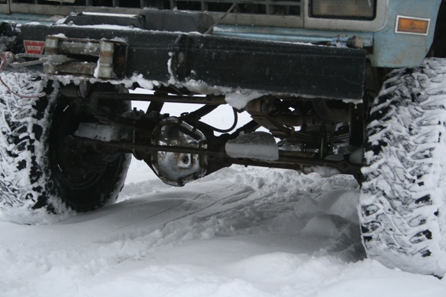 Front axle, suspension, and snow-covered tires of a light blue vehicle with a black bumper and winch